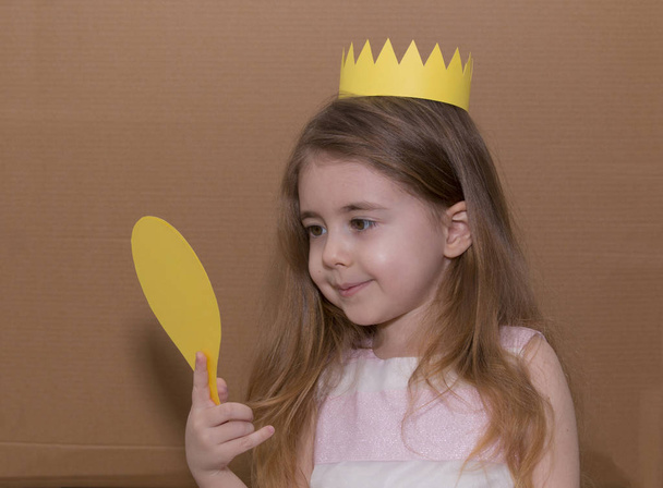 Portrait of emotional little girl with crown a yellow paper holding a mirror. handmade. cardboard background - Фото, изображение