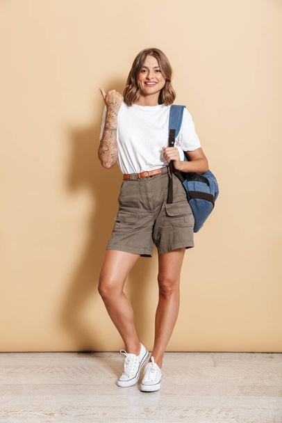 Image of content teen girl carrying backpack smiling and pointing finger isolated over beige background - Valokuva, kuva