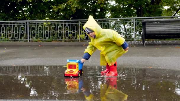Slow motion video of little toddler boy in rubber boots and raincoat plying with toy truck in big puddle after raining at park - Video
