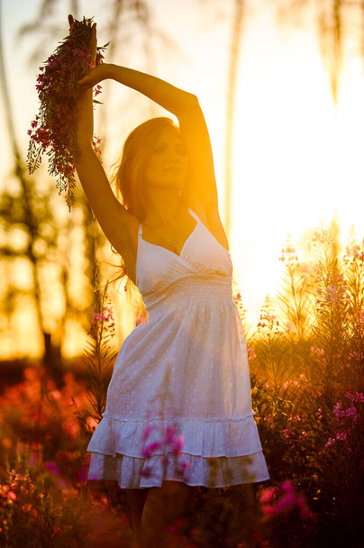 Joven chica rubia hermosa con una corona de flores recoge flores rosadas en el campo de floración de primavera al atardecer
. - Foto, imagen