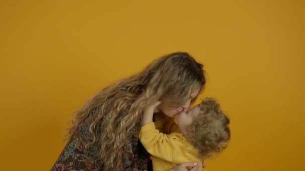Smiling mother kissing curly kid on yellow background - Záběry, video