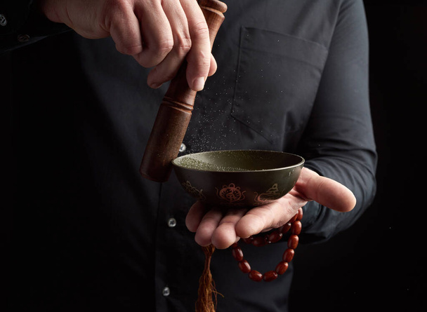Ένας ενήλικας με μαύρο πουκάμισο περιστρέφει ένα ξύλινο ραβδί γύρω από ένα χάλκινο θιβετιανό μπολ. τελετουργικό διαλογισμού, προσευχής και εμβάπτισης σε έκσταση. Εναλλακτική θεραπεία - Φωτογραφία, εικόνα