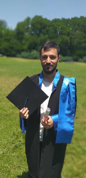 Young Graduate Person Holds His Diploma and Graduation Cap in a Grassy Field - Vertical Photo - Photo, Image