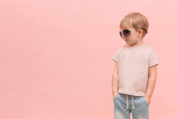 Un niño rubio de aspecto europeo está de pie con gafas de sol y se ve a la izquierda sobre un fondo rosa. Concepto con lugar para texto, para artículos sobre modelos, castings y desfiles de moda
. - Foto, Imagen
