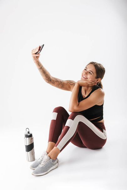 Image of joyful woman in sportswear taking selfie photo on cellphone after workout isolated over white background - Foto, Imagem