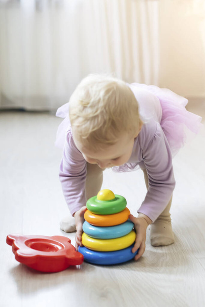 Mädchen trägt Spielzeugpyramide. Kind im Wohnzimmer sonnendurchflutet. Lernspielzeug für Kinder unter 2 Jahren. Buntes Spielzeug blau, gelb, rot, grün - Foto, Bild