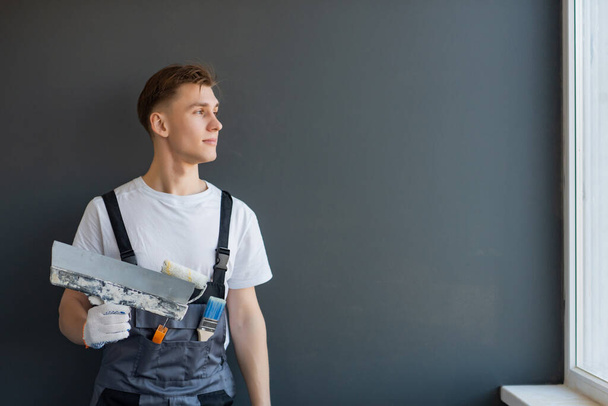 Young smiling male worker in overalls holds a spatula in his hand on a gray background - 写真・画像