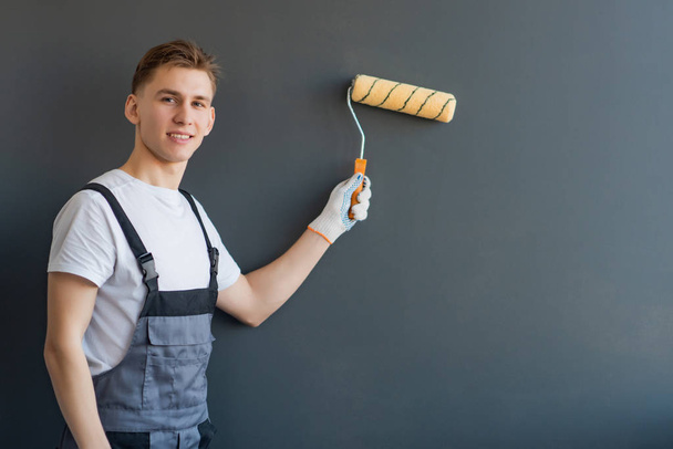 Portrait of a young smiling male worker in gray overalls and white t-shirt with a roller in his hand on a gray background. - Φωτογραφία, εικόνα