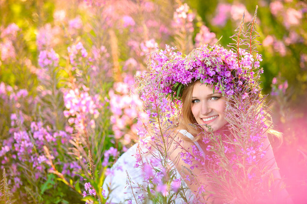 Young beautiful blonde girl with a flower wreath on her head gathers pink flowers in the spring blooming field at sunset. - Foto, afbeelding