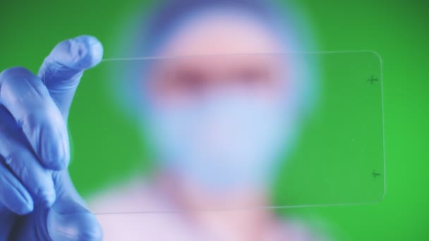 πράσινο φόντο. close-up, γιατρός ντυμένος με ιατρικό καπέλο, μάσκα, μπλε ιατρικά γάντια, κρατά μια γυάλινη κάρτα και κλικ σε αυτό. δυνατότητα τοποθέτησης διαφήμισης, κειμένου ή βίντεο στην κάρτα. - Πλάνα, βίντεο