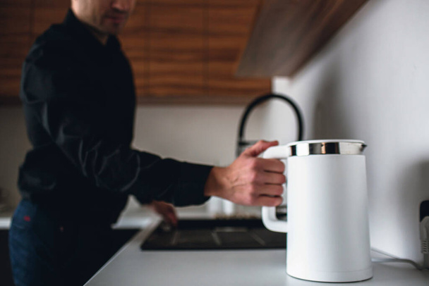 Young businessman in black shirt stand in kitchen and hold white electric smart kettle. Guy want to heat up water. Hold big finger on button. Ready to heat water. Coffee break. - Fotografie, Obrázek