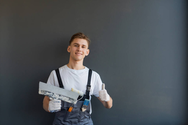 Young smiling male worker in overalls holds a spatula in his hand on a gray background - Фото, зображення
