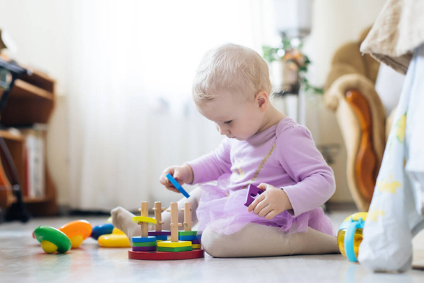 dívka hraje hračky v obýváku. Montessori dřevěná hračka skládaná pyramida. Kruh, quadra, trojúhelník, obdélník dřevěné prvky dětských hraček. Barevná hračka modrá, žlutá, červená, zelená. - Fotografie, Obrázek