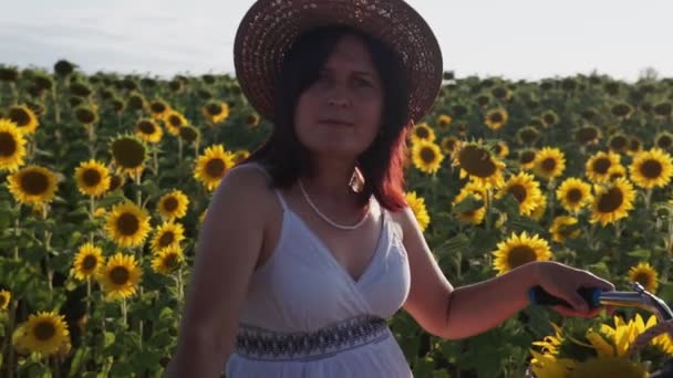 Une femme avec un chapeau et une robe blanche avec un vélo marche à travers le pollen avec des tournesols - Séquence, vidéo
