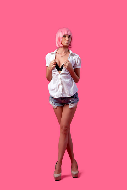 Attractive slim woman classy posing in a white shirt, short denim shorts and a pink wig with a hairstyle on a white isolated background. Woman posing in a photo studio. Bright summer design concept. Woman in a bright wig - Fotoğraf, Görsel