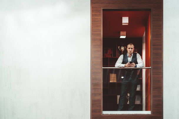 Empresário hispânico bonito grave está encostado contra corrimão varanda enquanto usa seu smartphone em um recesso de uma área de chillout escritório moderno, com um espaço de cópia à esquerda para uma mensagem
 - Foto, Imagem