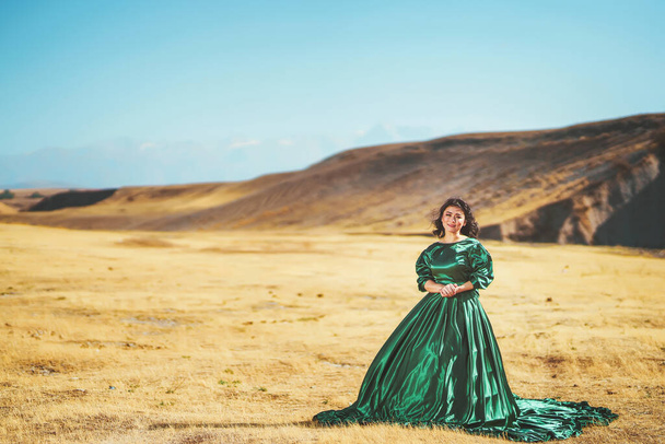 Woman in green dress on the background of mountains in south Kazakhstan. Young woman in  fairy tale ball gown on background of mountains and yellow steppe. Beautiful girl in long dress in mountains - Photo, Image