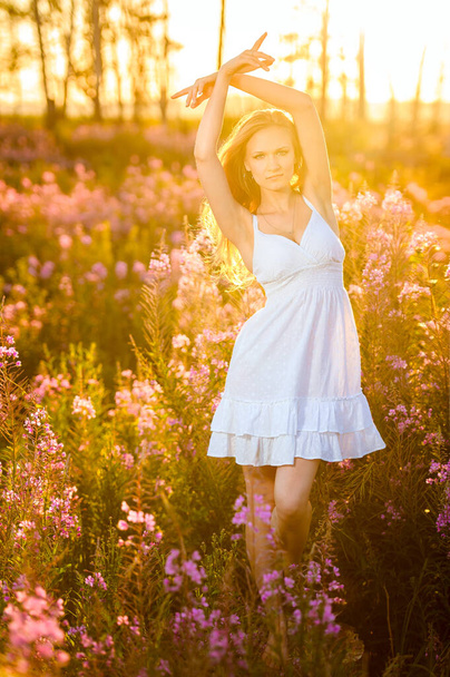 Молодая красивая блондинка с цветочным венком собирает розовые цветы весной цветущие поля на закате
. - Фото, изображение