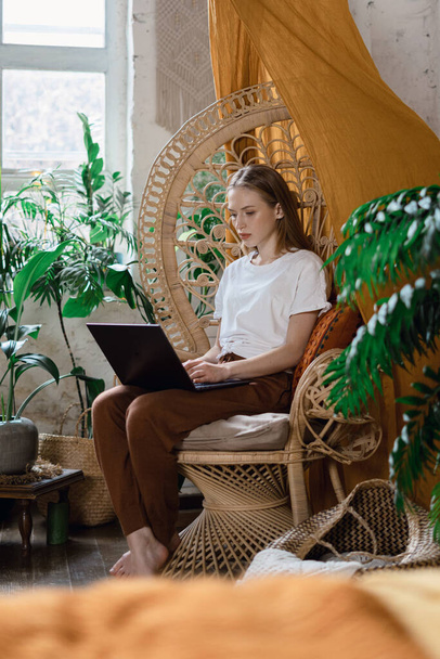 Вертикальное фото молодой взрослой женщины, создающей онлайн контент для блога, сидящей в плетеном кресле дома, используя современный ноутбук
 - Фото, изображение
