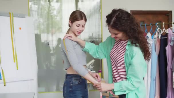 Joven costurera atractiva tomar medidas de chica bonita con cinta centímetro en acogedor estudio de sastrería
  - Metraje, vídeo