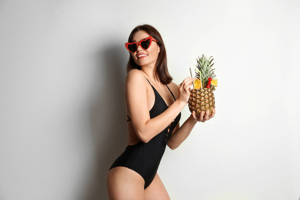 Beautiful woman in stylish swimsuit holding tropical cocktail on light grey background - Photo, image