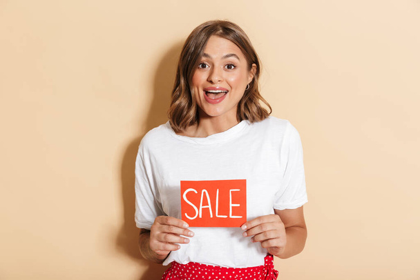 Cheerful young girl wearing casual outfit standing isolated over beige background, showing sale banner - Foto, imagen