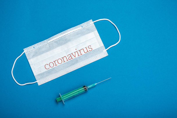 Green syringe and surgical mask with rubber ear straps. Typical 3-ply surgical mask to cover the mouth and nose. Procedure mask from bacteria. Protection concept. On blue background. - 写真・画像