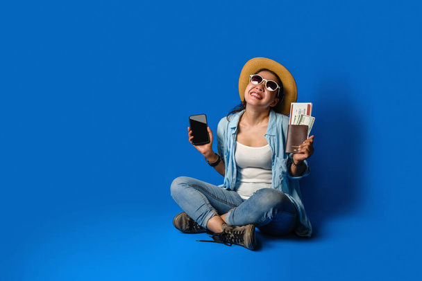 Menina viajante em um vestido azul está sorrindo feliz com um rosto alegre e segurando passaporte com segurando telefones celulares na mão, no fundo da cor azul. conceito de viagem - Foto, Imagem