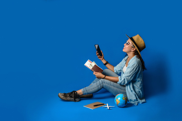 Νεαρός ασιάτης ταξιδιώτης χαρούμενος με μπλε πουκάμισο κρατώντας διαβατήριο με τα κινητά τηλέφωνα στο χέρι. έννοια ταξίδια με εξοπλισμό για τους ταξιδιώτες διακοπές, σε μπλε χρώμα φόντο. Ταξίδια χαλαρώστε - Φωτογραφία, εικόνα