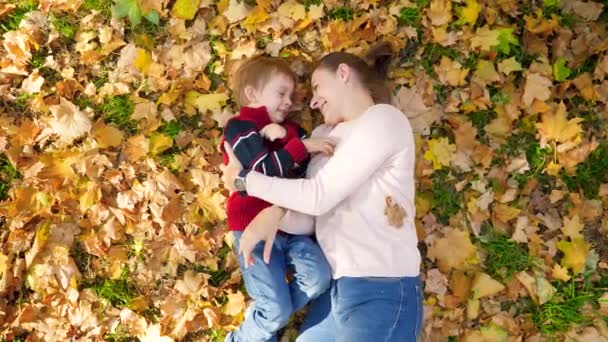 4k vídeo de menino com a mãe brincando, fazendo cócegas entre si, abraçando e beijando enquanto deitado em folhas amarelas e grama no parque de outono
 - Filmagem, Vídeo