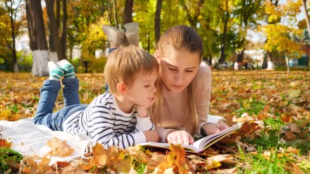 4k video de la joven madre con su pequeño hijo acostado en la manta en el parque de otoño y libro de lectura
 - Metraje, vídeo