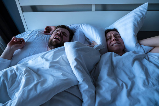 Nešťastná žena nemůže usnout, zakryje si uši polštářem, protože její manžel chrápe v posteli. Koncept ženské nespavosti - Fotografie, Obrázek