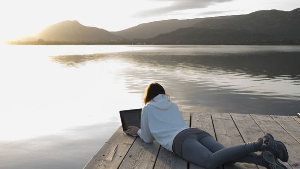 Internet freelance job choice concept: een jonge vrouw werkt op haar laptop liggend op een pier bij een meer bij zonsondergang - Foto, afbeelding