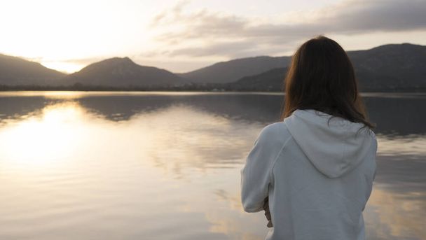 Mujer joven pensativa con el pelo largo visto por detrás mira el lago al atardecer
 - Foto, Imagen