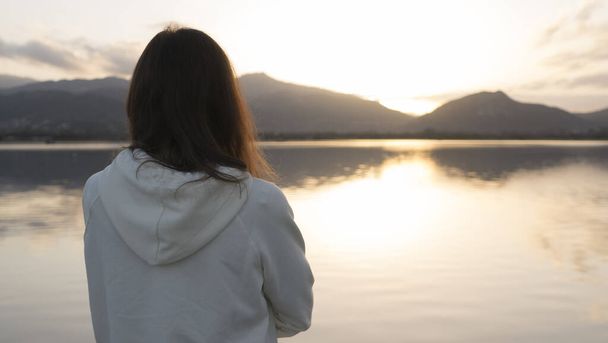 Eine junge Frau mit langen Haaren blickt von hinten auf den See bei Sonnenuntergang - Foto, Bild