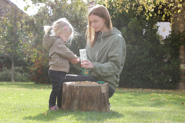 Mother and her toddler girl putting seeds for birds in a bird feeder. Quality outdoor family time together. Encouraging wildlife in the garden with bird feeder. - Photo, Image