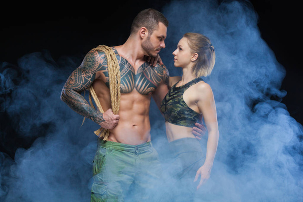 Hombre musculoso sin camisa con tatuaje y cuerda posando con hermosa deportista en humo sobre fondo negro
 - Foto, Imagen