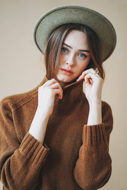 Fiatal gyönyörű hosszú barna hajú lány kék szeme filc kalap és barna kötött pulóver nézi kamera bézs háttér - Fotó, kép