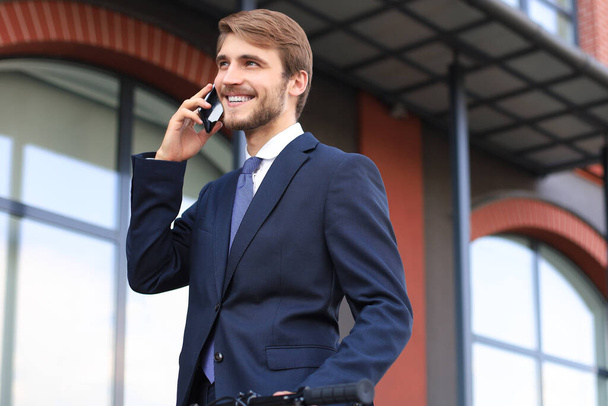 Портрет усміхненого молодого бізнесмена, одягненого в костюм, який говорить на мобільному стоячому на відкритому повітрі
 - Фото, зображення