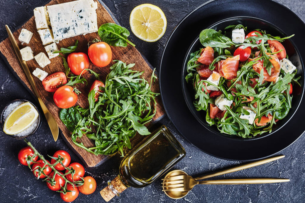 close-up of delicious healthy salmon arugula blue cheese tomato salad in a black bowl with golden cutlery and ingredients, horizontal view from above, flat lay - Fotografie, Obrázek