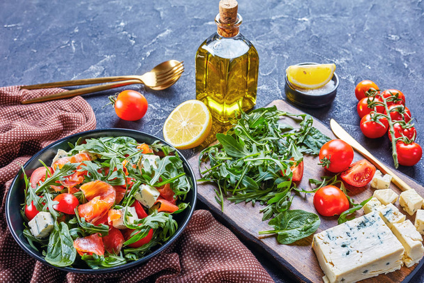 close-up of delicious healthy salmon arugula blue cheese tomato salad in a black bowl with golden cutlery and ingredients, horizontal view from above,  free space - Fotografie, Obrázek