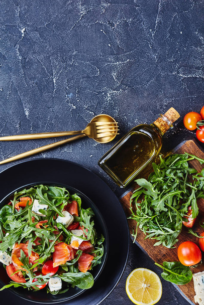 close-up of delicious healthy salmon arugula blue cheese tomato salad in a black bowl with golden cutlery and ingredients, vertical view from above, flat lay, free space - Photo, Image
