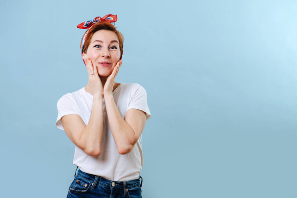 Portrait of a cheerful middle aged housewife with short ginger hair and colorful headband making surprise gesture. She's wearing casual clothes: striped T-shirt and jeans. Over blue background. - Fotografie, Obrázek