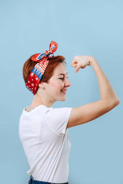 Portrait of a cheerful middle aged housewife with short ginger hair and colorful headband tencing biceps, showing off her strength. She's wearing casual clothes: white T-shirt and jeans. Over blue - Φωτογραφία, εικόνα