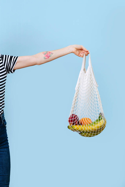 Mano de mujer sosteniendo una bolsa de red con frutas sobre fondo azul. Cortado, sin cabeza
. - Foto, Imagen