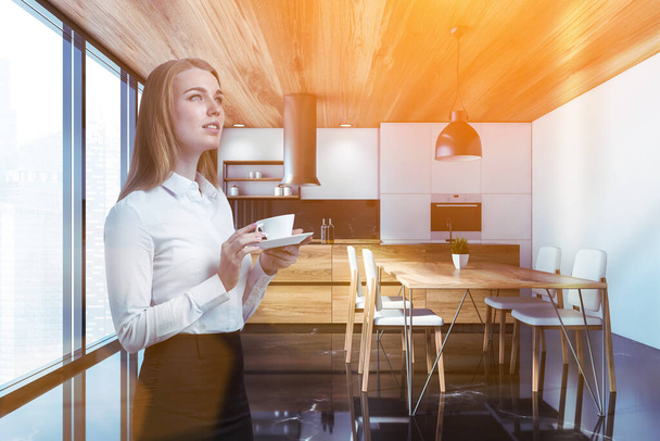 Beautiful blonde woman with coffee standing in luxury kitchen with white walls, black marble floor, wooden island and dining table with white chairs. Toned image - Valokuva, kuva