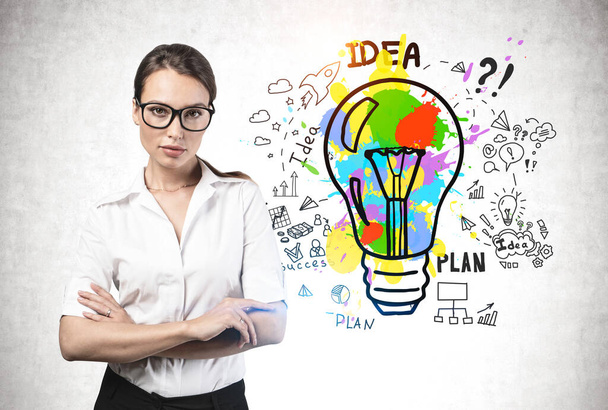 Confident young woman in glasses standing with crossed arms near concrete wall with colorful business idea sketch. Concept of creativity and startup - Φωτογραφία, εικόνα
