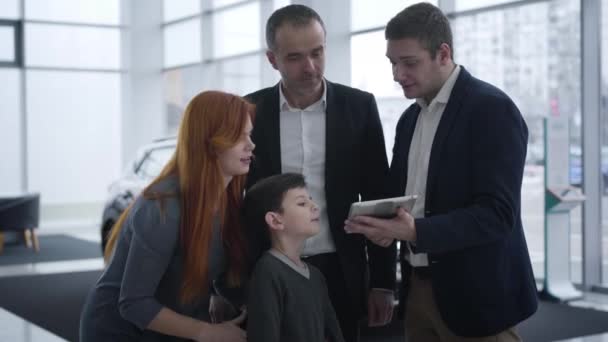 Caucasian family talking with car dealer in showroom. Husband, wife and little boy choosing new vehicle in dealership. Mother raising son and showing him dealers tablet. Auto industry, buying. - Video, Çekim