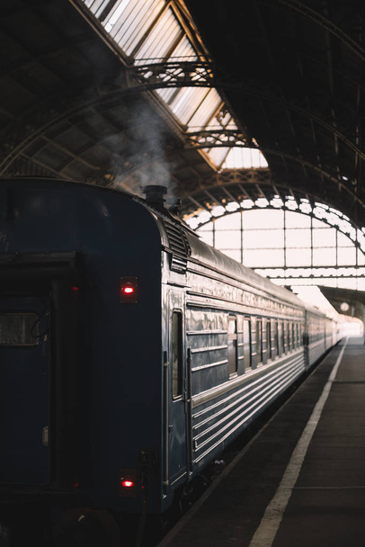 Retro passenger railway wagon preparing to depart from the covered platform. Smoke escapes from the chimney. Exterior of vintage railcar of train. Vertical photo. Vacation and tourism concept. - Фото, зображення