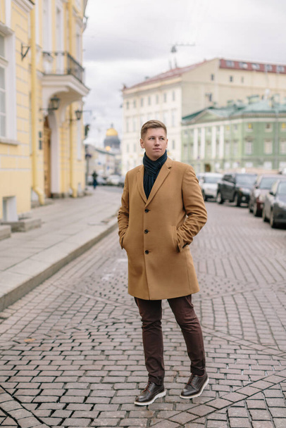 Attractive blond caucasian man in beige coat, with dark blue scarf tied around his neck, brown pants and dark leather shoes keeps hands in pockets. Selective focus on pretty hipster. Lifestyle concept - Фото, зображення